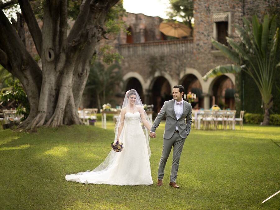 Una pareja tomados de la mano en una zona cubierta de hierba, con un gran árbol y un edificio con arcos al fondo.