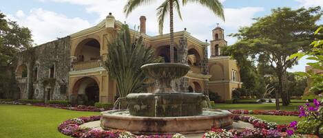 Un edificio histórico con una fuente central rodeada de un jardín cuidado y palmeras.