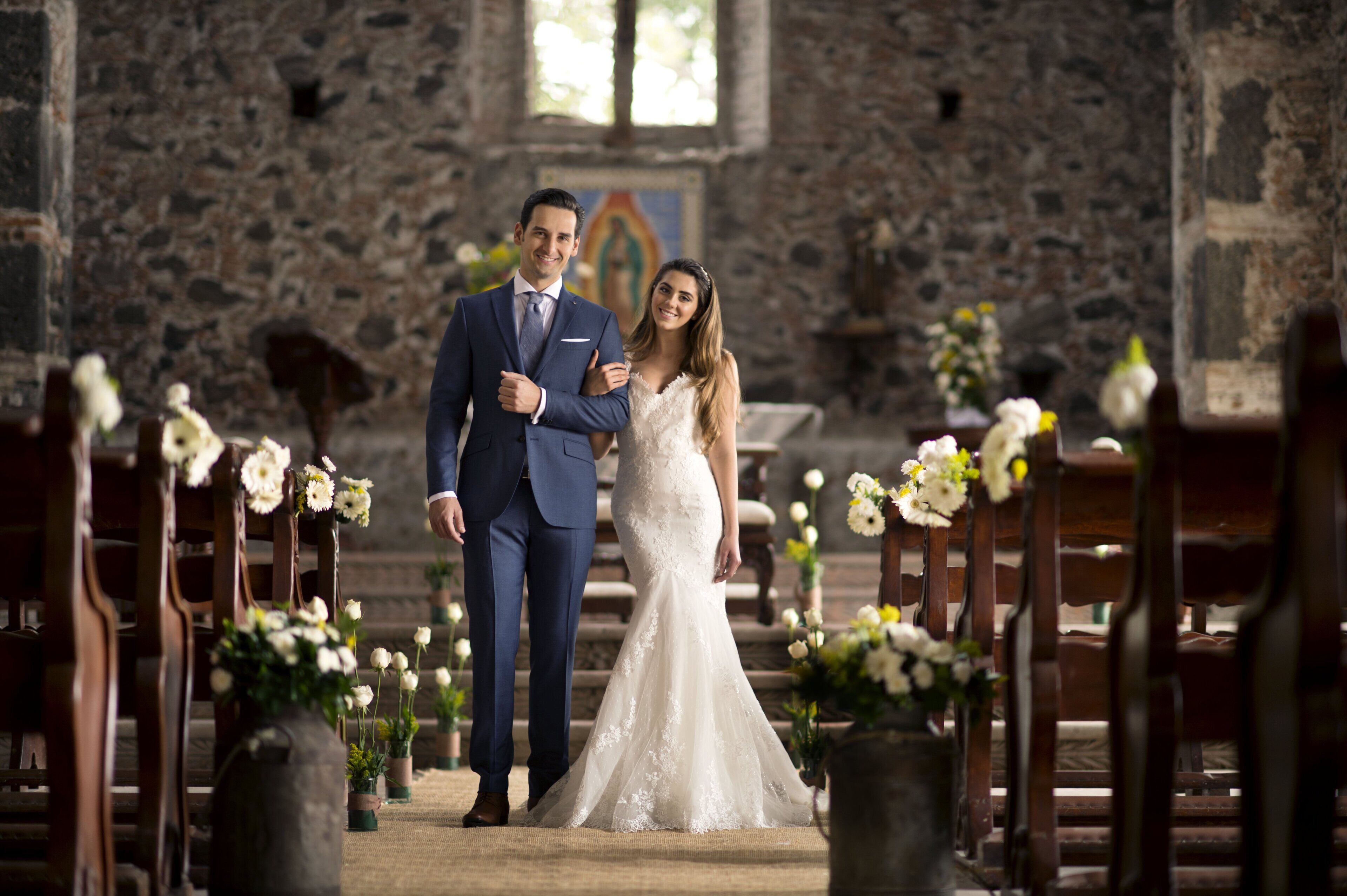 Una pareja vestida de novios caminando por el pasillo de una iglesia, con bancas a ambos lados.