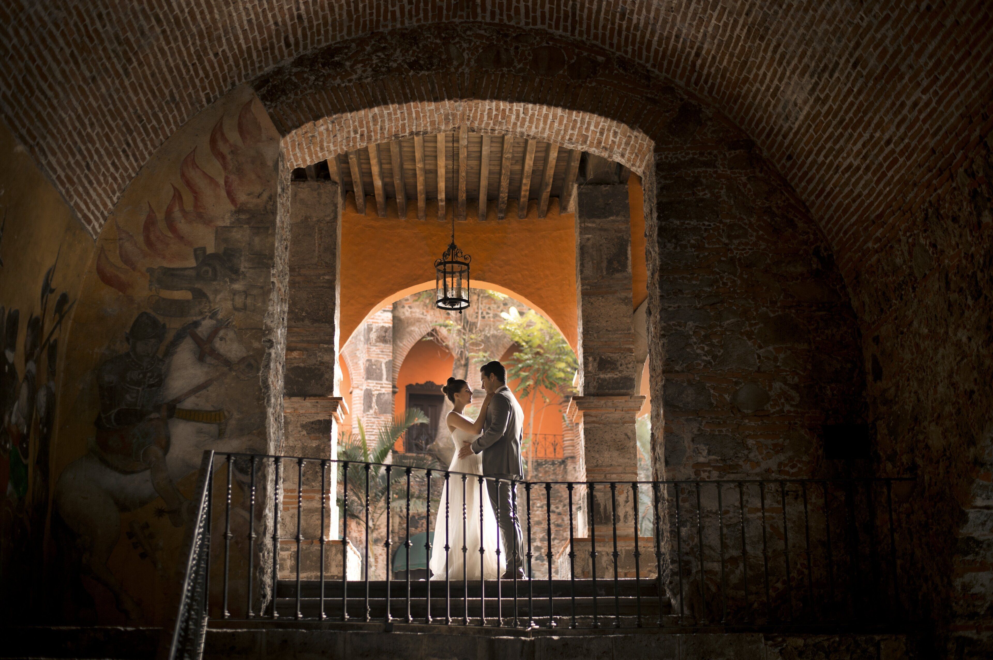 Una pareja parada en un corredor de piedra con arco, barandilla de hierro forjado, un farol colgante y un mural en la pared.