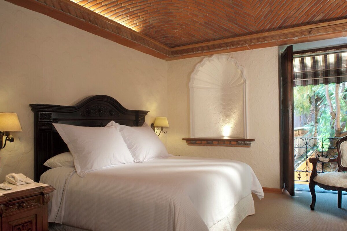 Habitación de hotel con una cama grande, dos lámparas de noche, una mesita de noche con teléfono y vistas a la vegetación a través de una ventana con balcón de hierro forjado.