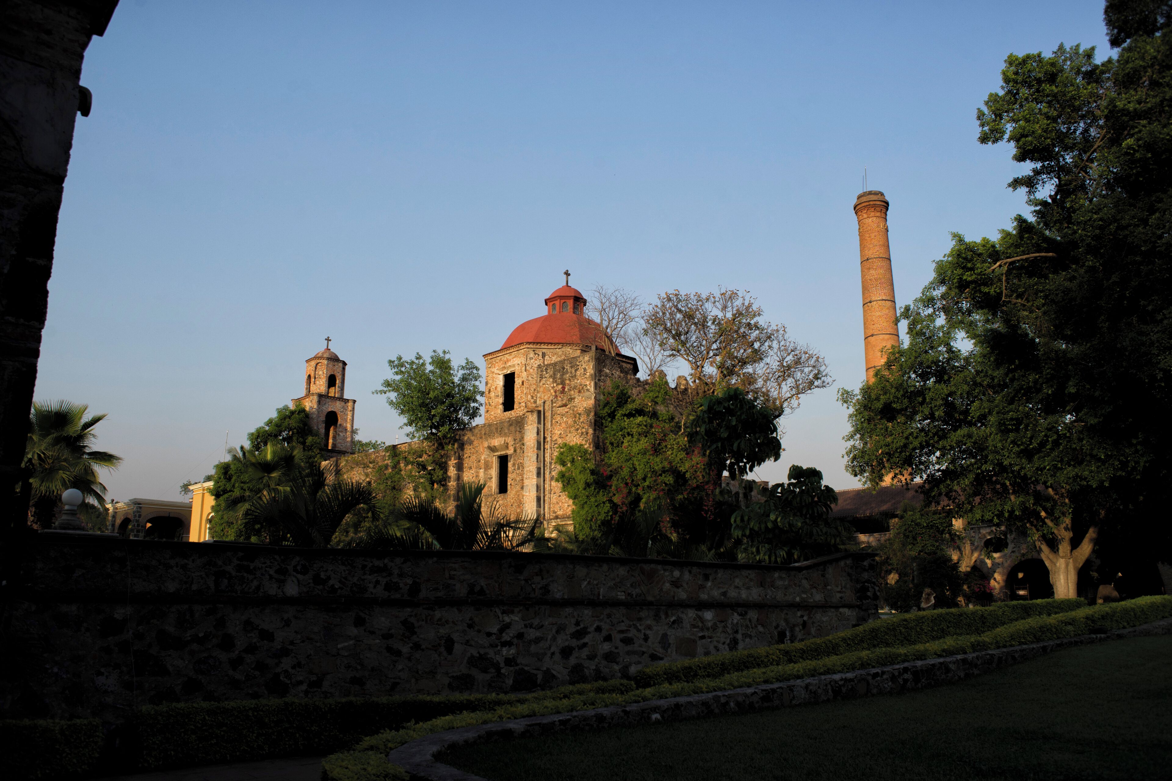 A historic building with a red dome and a bell tower, surrounded by trees and a stone wall.