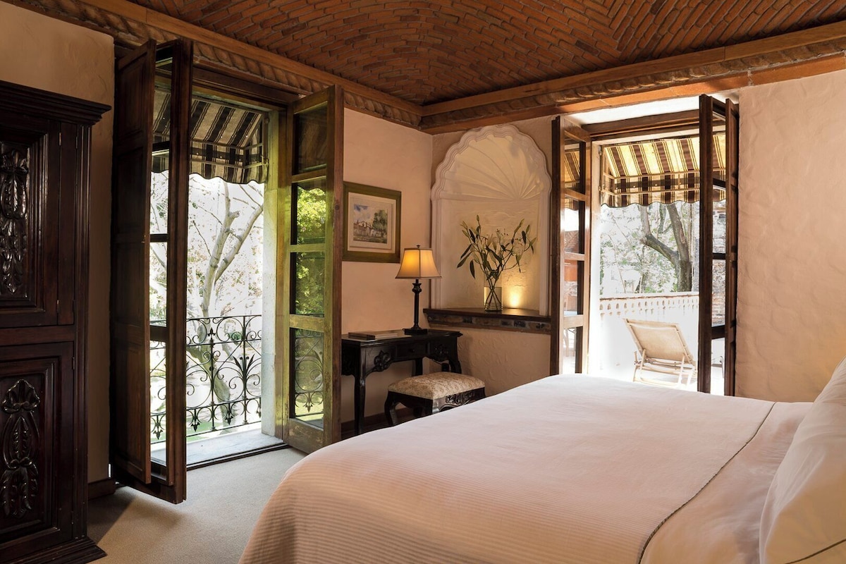 Un dormitorio con una cama grande, un escritorio con lámpara, y una silla. Hay una ventana con vistas a árboles y un balcón.