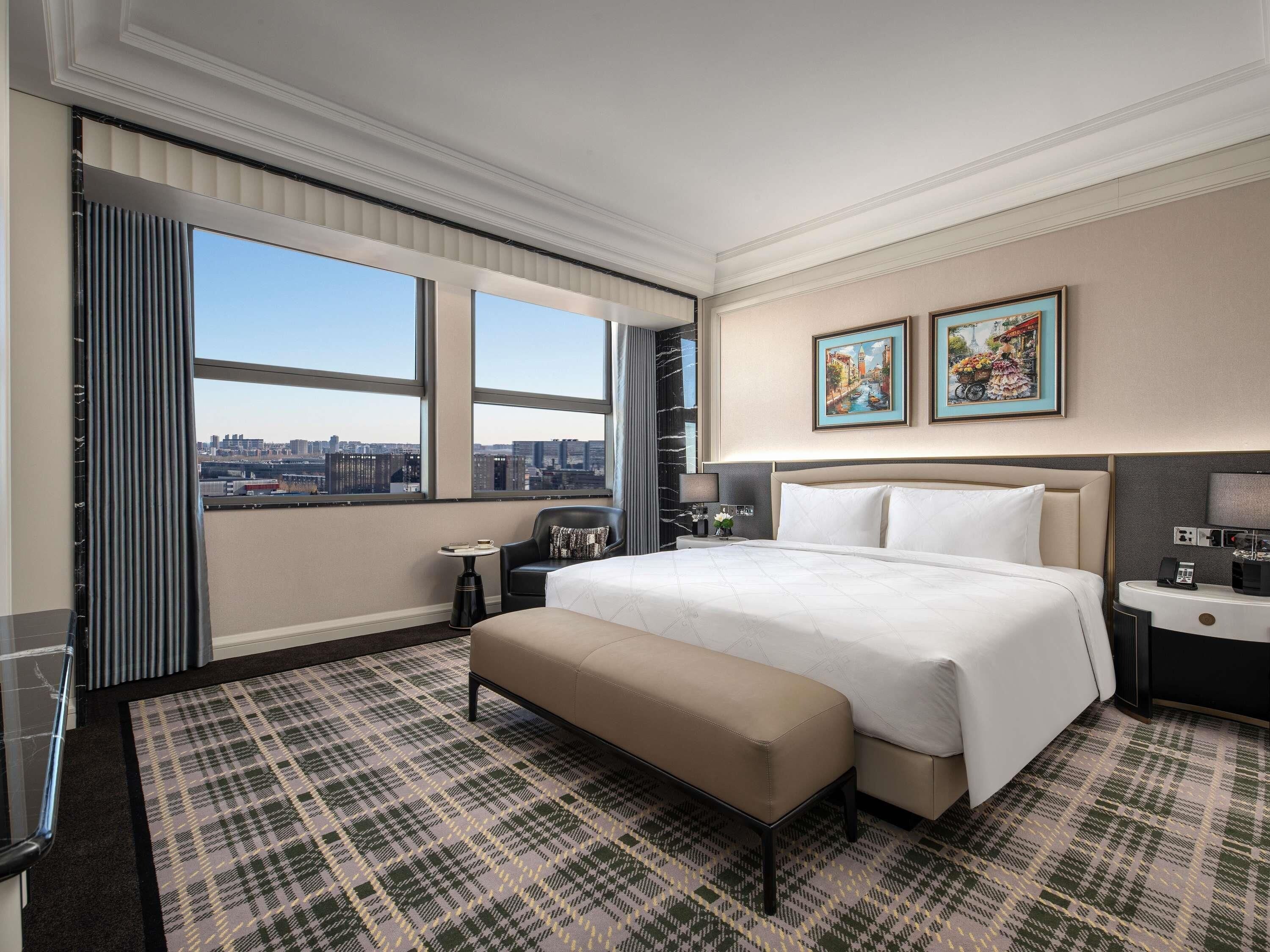 A hotel room with a large bed, a bench, a small table, a chair, and a city view through large windows.