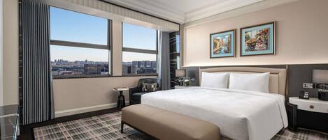 A hotel room with a large bed, a bench, a small table, a chair, and a city view through large windows.