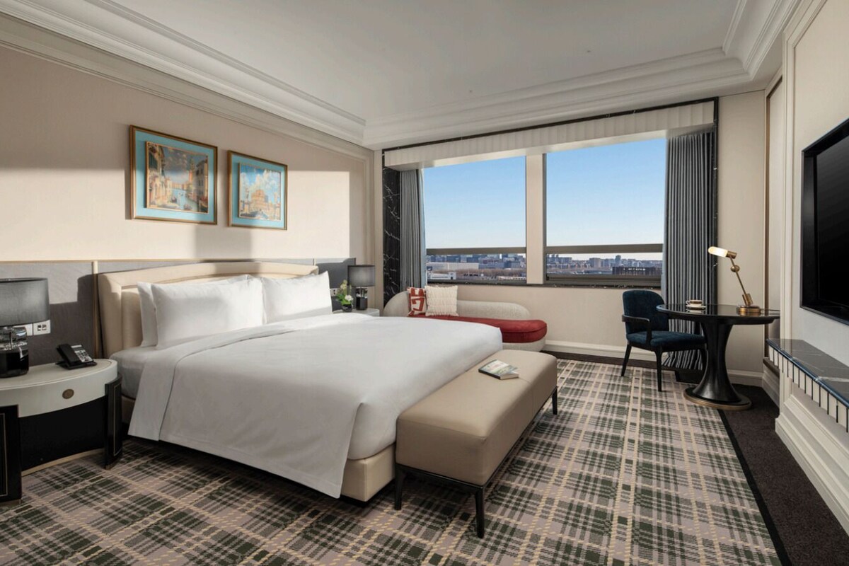 A hotel room with a large bed, a desk with a lamp, a TV, a sofa, and a view of the city.