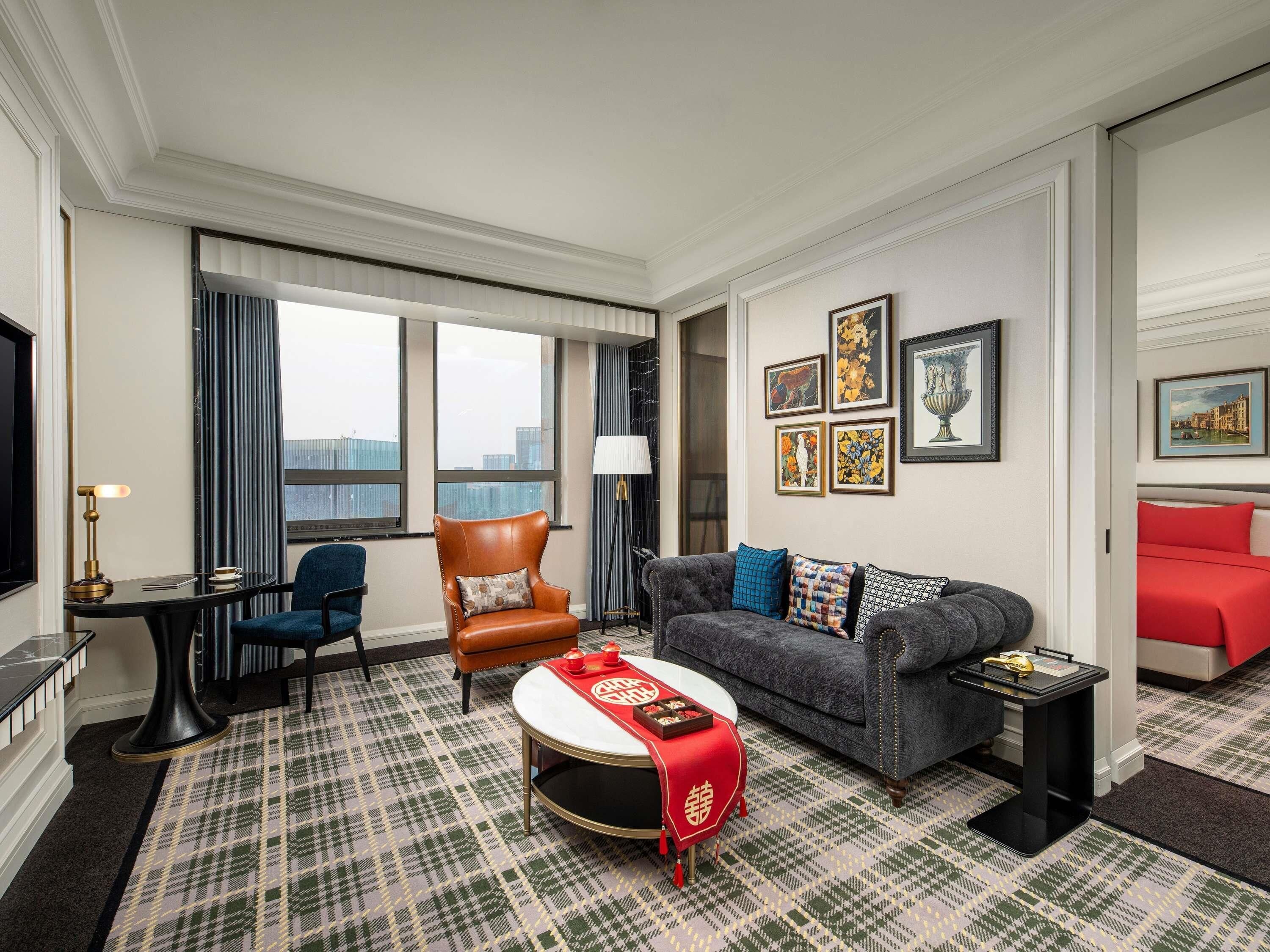 A hotel room with a large window, a sofa, a chair, a small table, and a red bed.