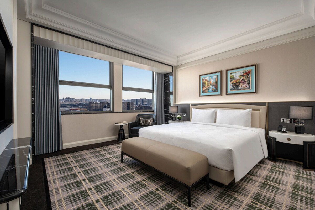 A hotel room with a large bed, a bench, a desk, a chair, a TV, and a city view.
