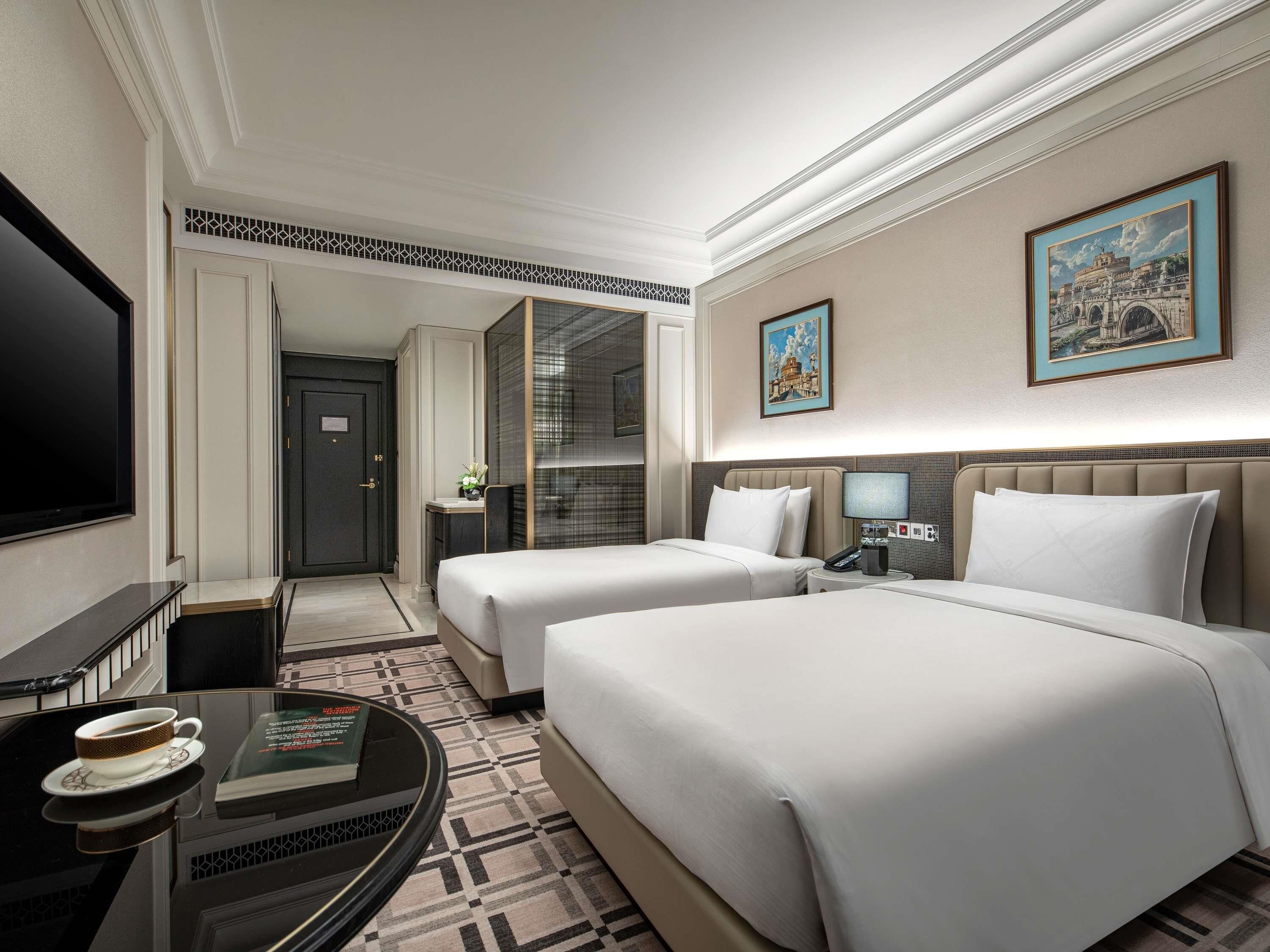 A hotel room with two beds, a TV, a desk with a cup and book, and a patterned floor.