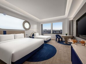 A hotel room with a large bed, a desk, a chair, a round table, a TV, and a view of the city.