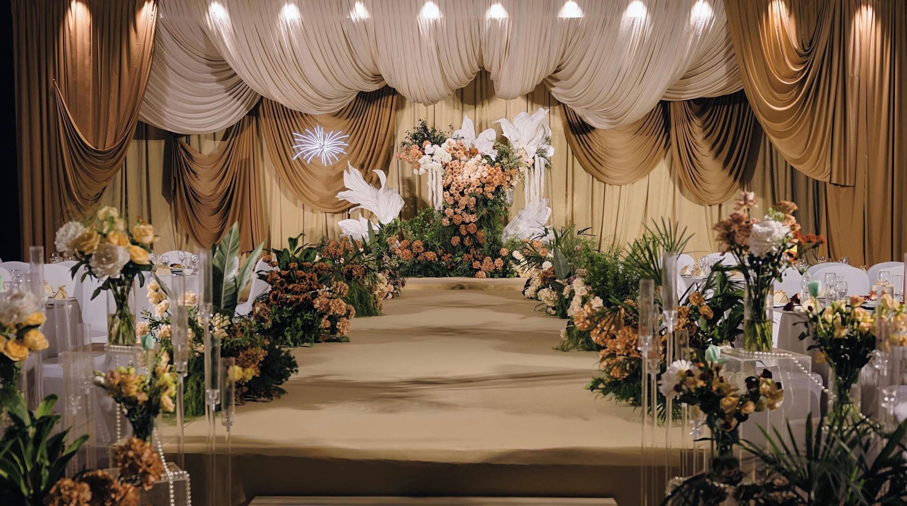 An indoor venue with a stage, draped curtains, and floral arrangements.