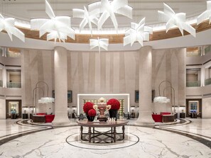 A spacious lobby with a central round table, high ceilings with unique lighting fixtures, and a grand staircase.