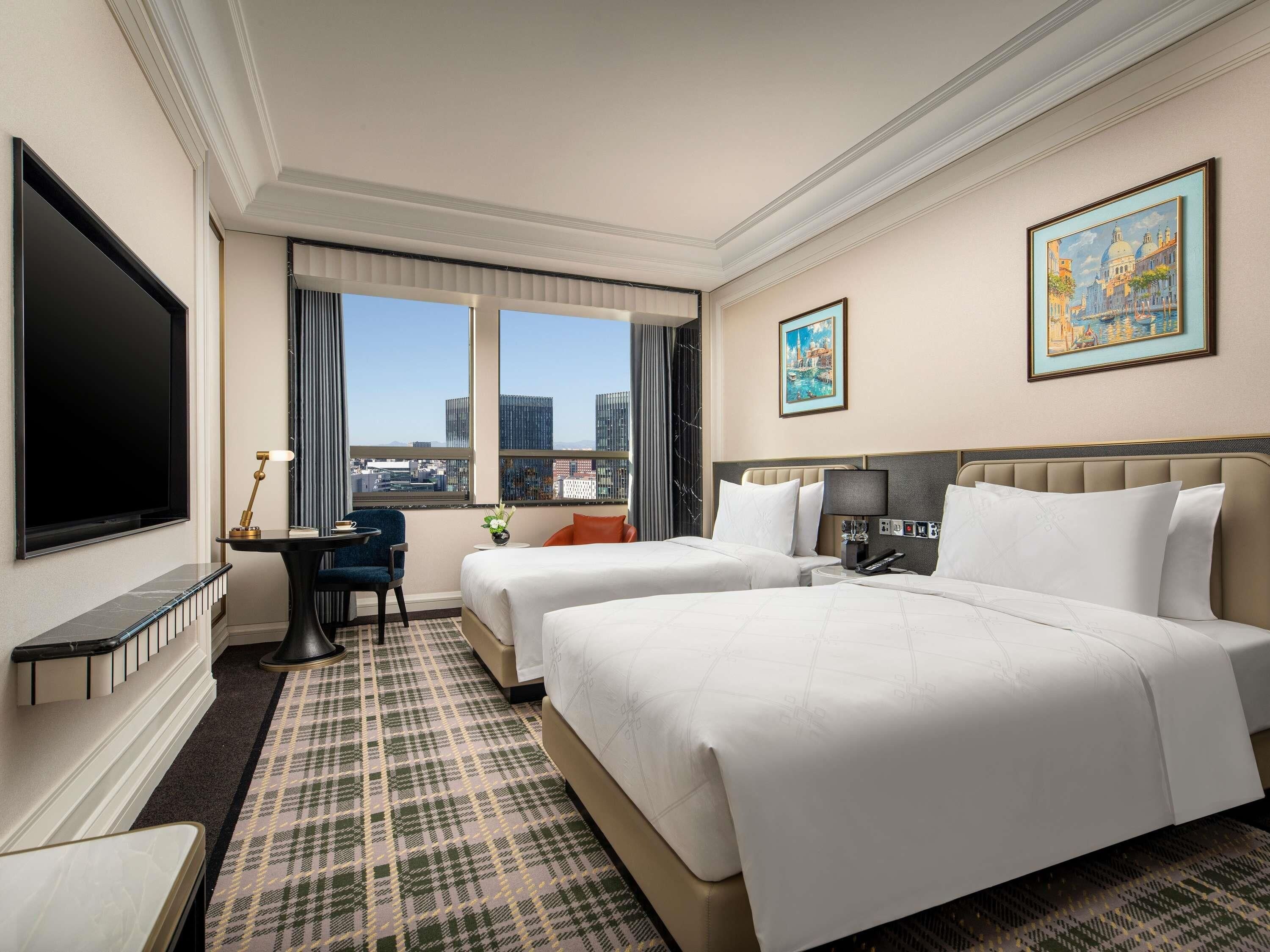 Habitación de hotel con dos camas, televisión, escritorio y vista a la ciudad.