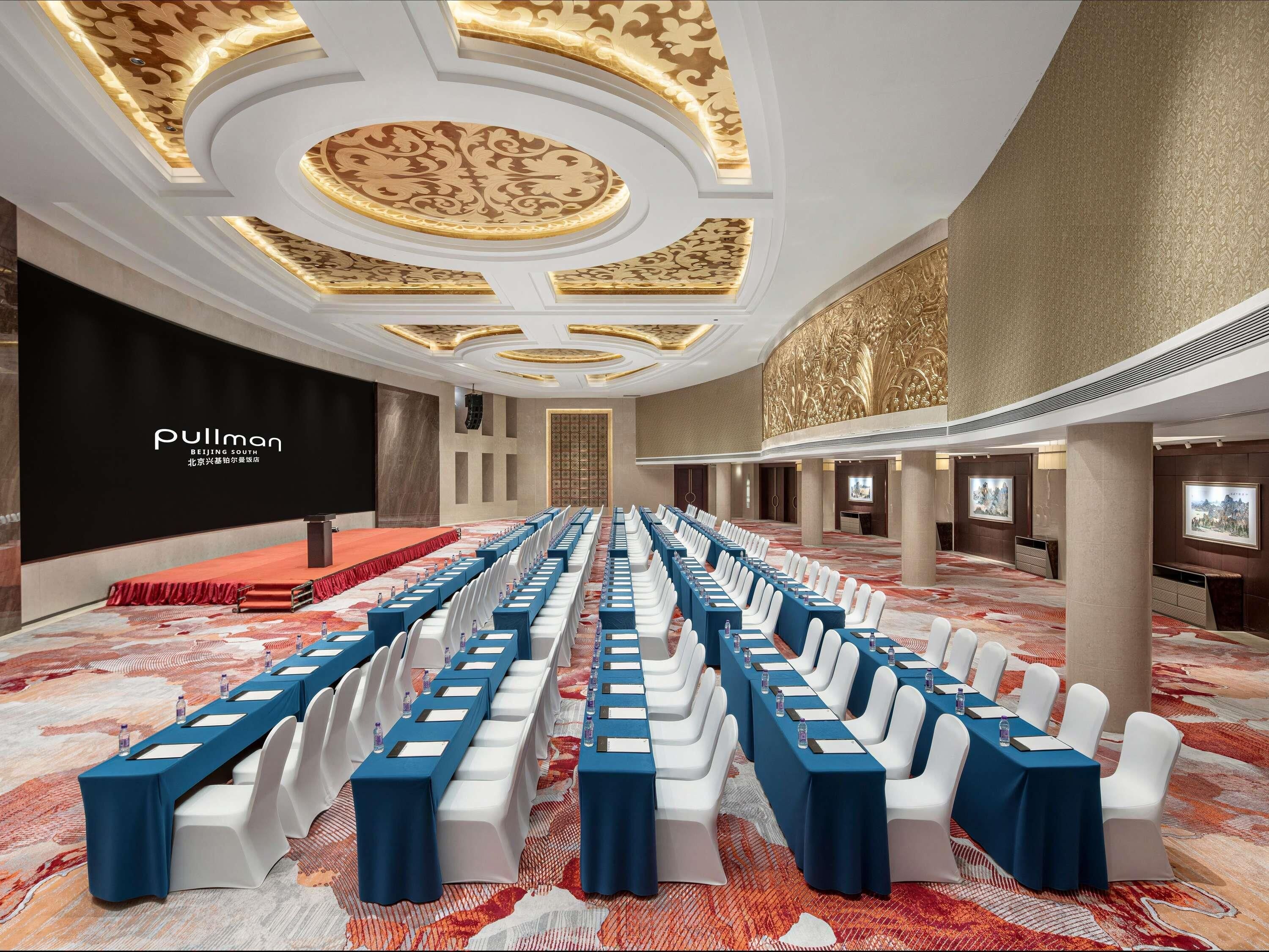 A conference room with rows of white chairs, blue tables, and a stage with a red carpet.