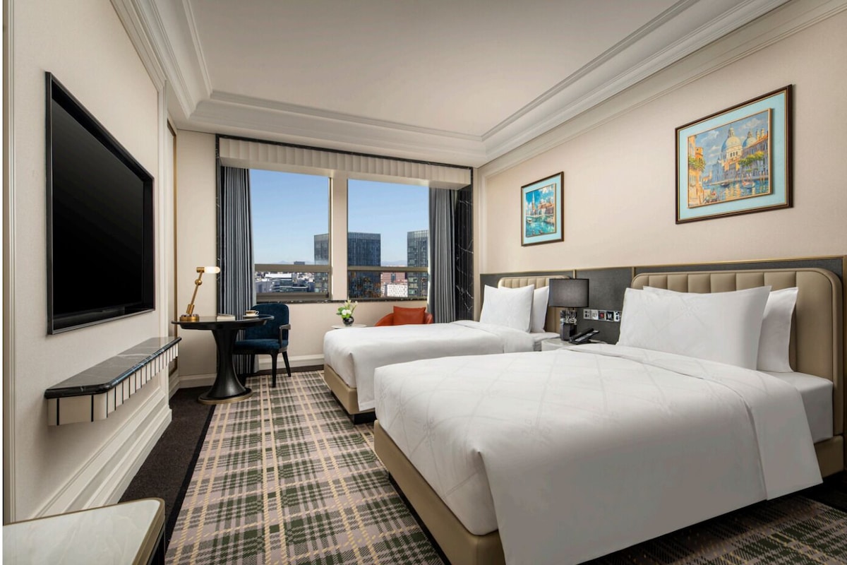 A hotel room with two beds, a TV, a desk, and a city view.