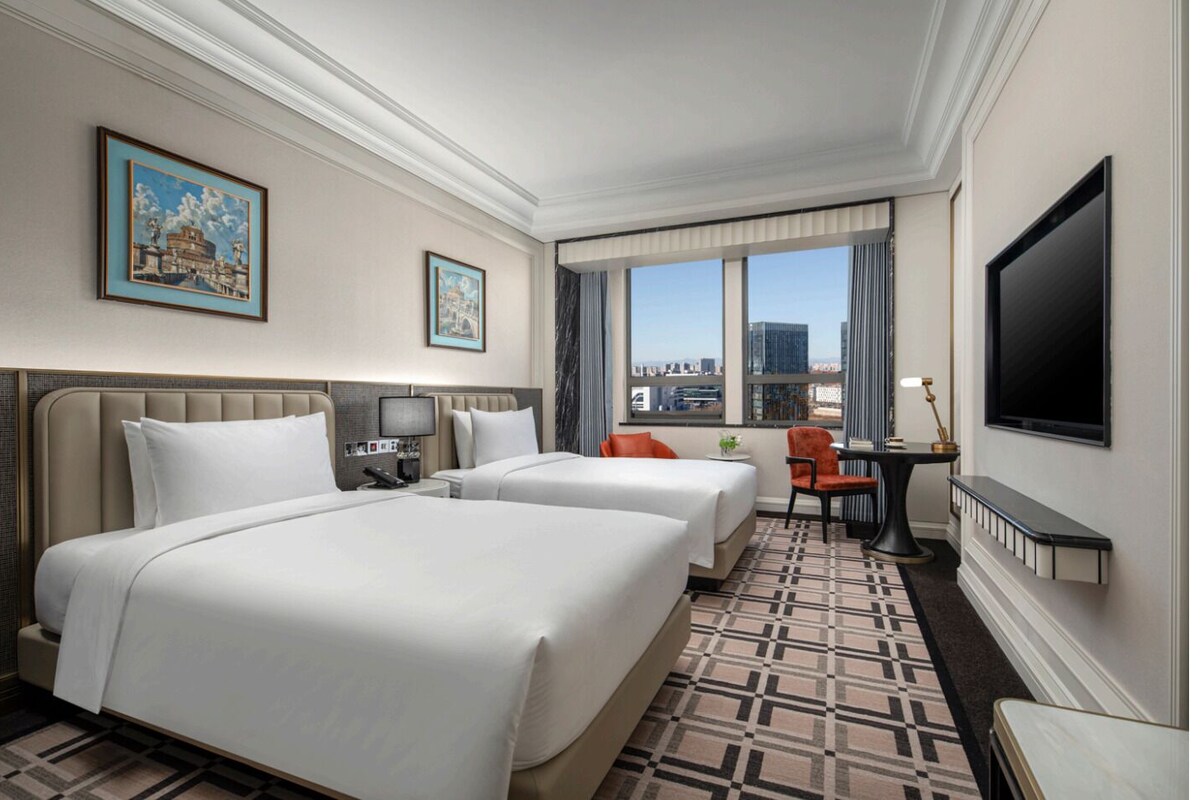 A hotel room with two beds, a desk, a chair, a TV, and a city view.