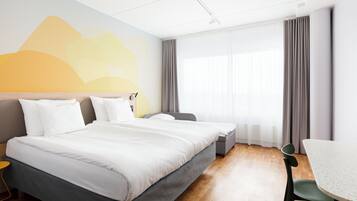 A hotel room with a large bed, a desk, and a chair. There is a window with curtains, a wall with a mountain design, and a ceiling with recessed lighting.