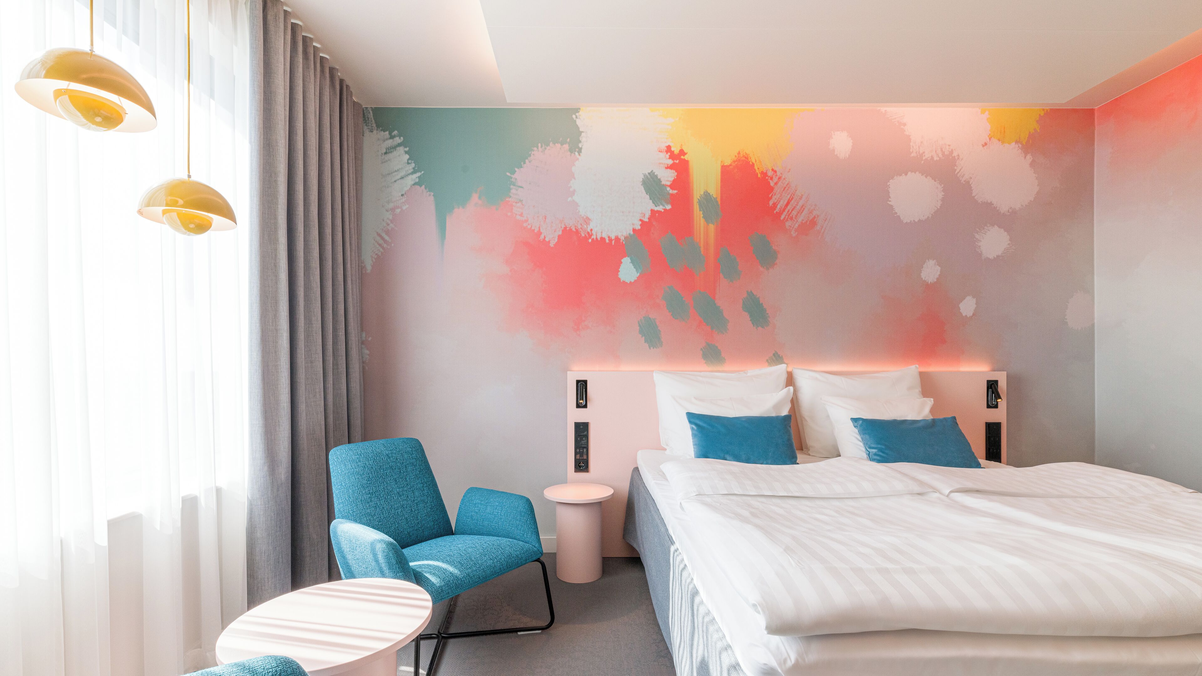 Una habitación de hotel moderna con una cama, un sillón azul, una mesita redonda y un mural abstracto de colores en la pared.