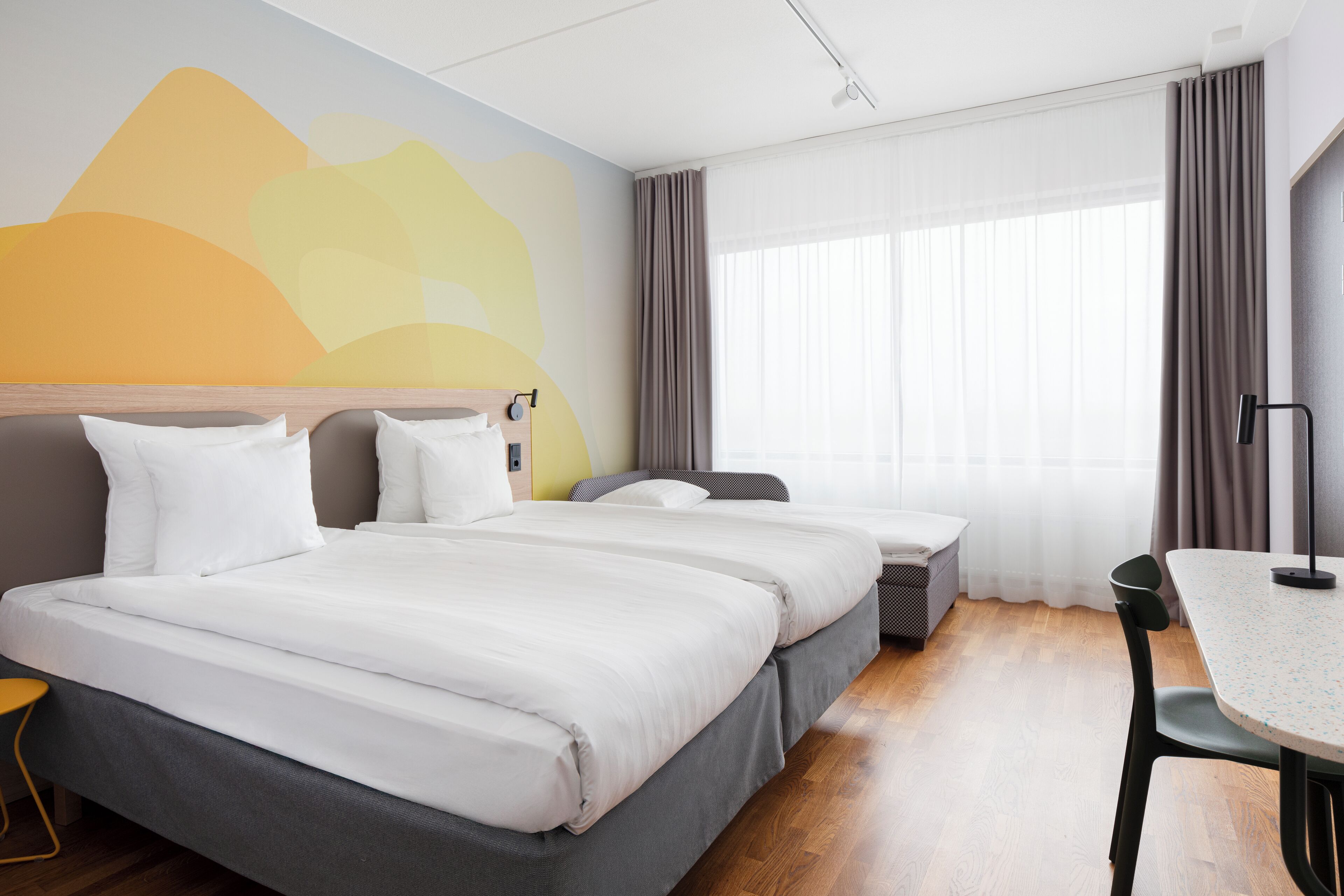Quarto de hotel com uma cama grande, uma mesa pequena e uma cadeira. Há uma janela com cortinas e uma parede com um design abstrato colorido.