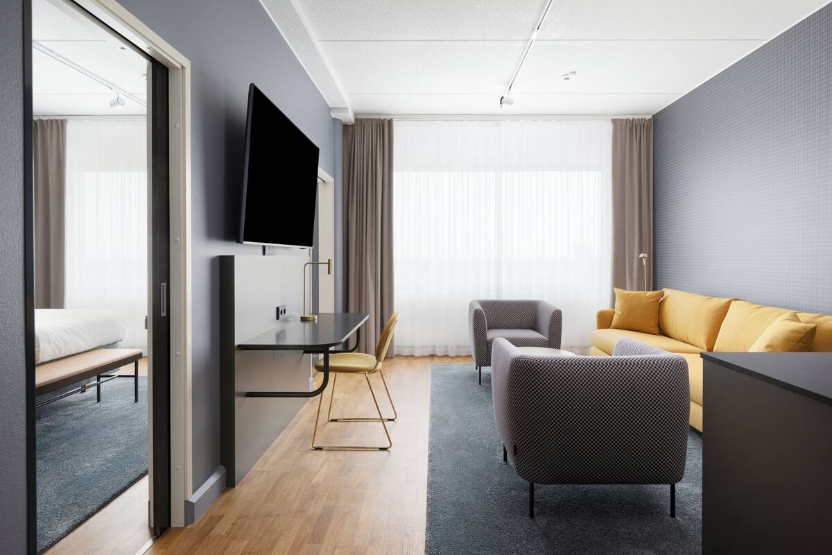 A modern hotel room with a bed, a desk, a TV, two armchairs, and a sofa.