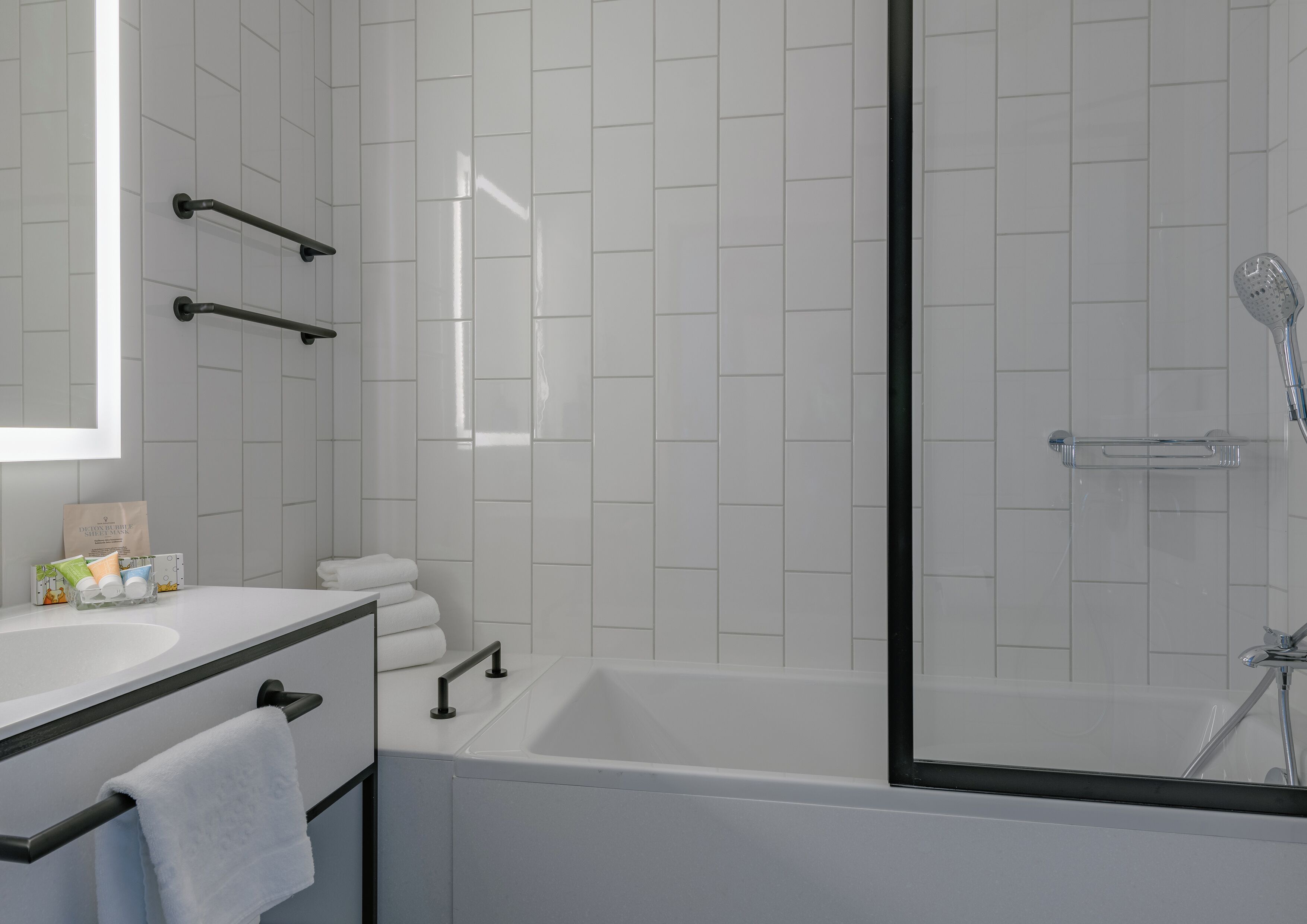 Ein modernes Badezimmer mit weiß gefliester Wand, einer Badewanne, einer Dusche und einem Waschtisch mit Spiegel und Handtuchhalter.
