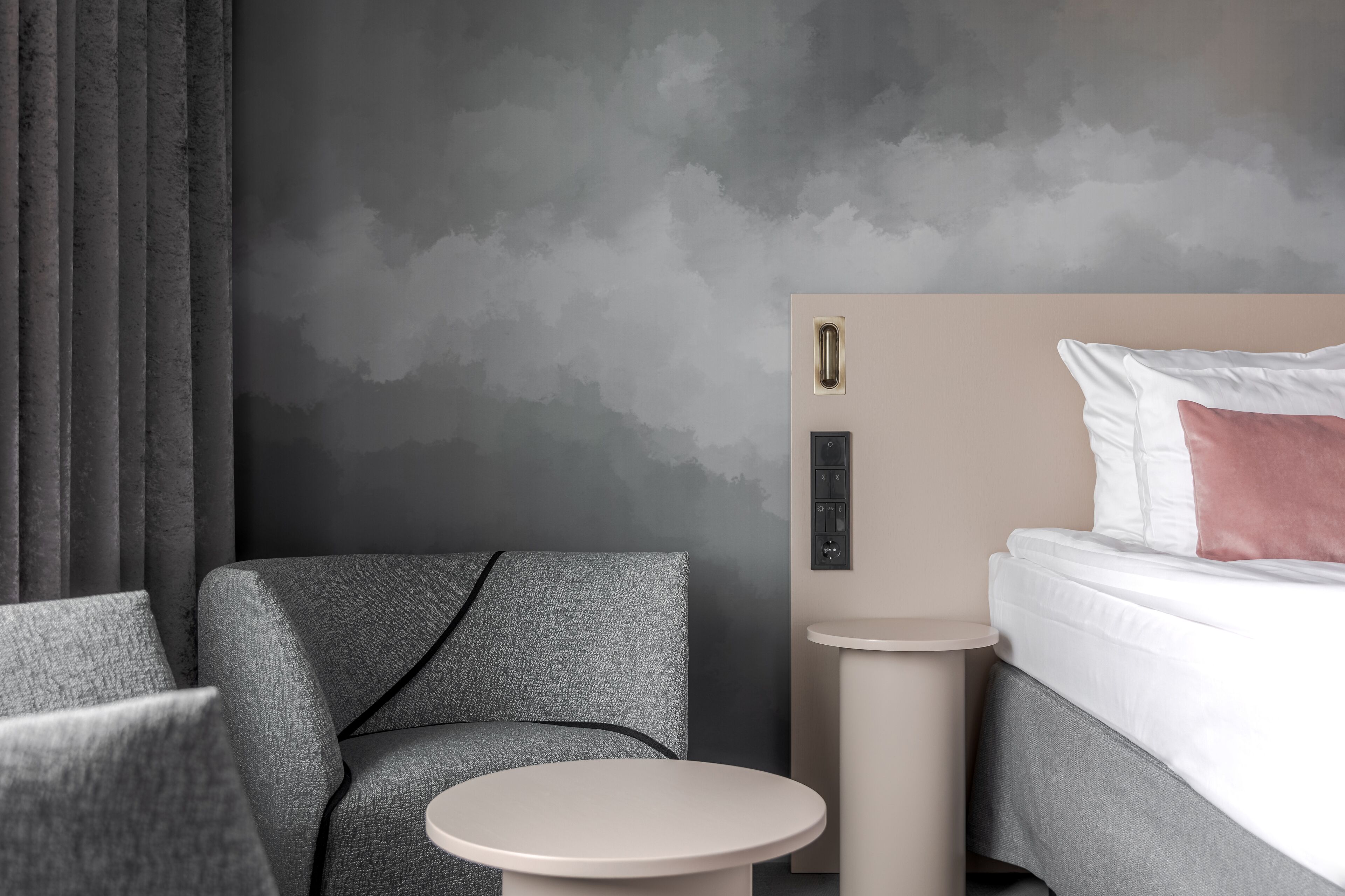 A modern hotel room with a grey sofa, a round table, a bed with white and pink pillows, and a grey textured wall.