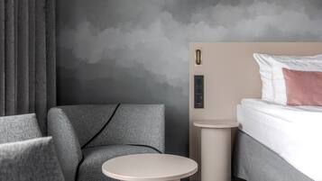 A modern hotel room with a grey sofa, a round table, a bed with white and pink pillows, and a grey textured wall.
