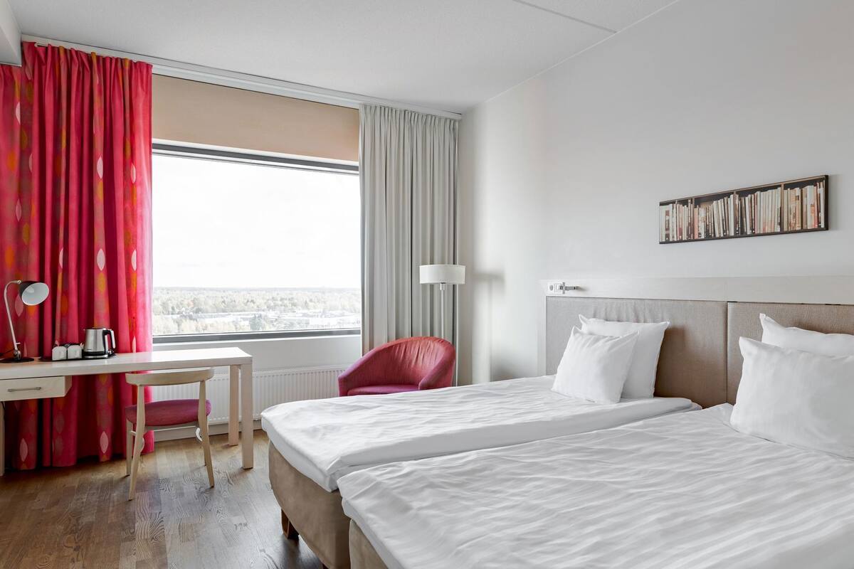 A hotel room with two beds, a desk, a chair, a lamp, and a bookshelf.