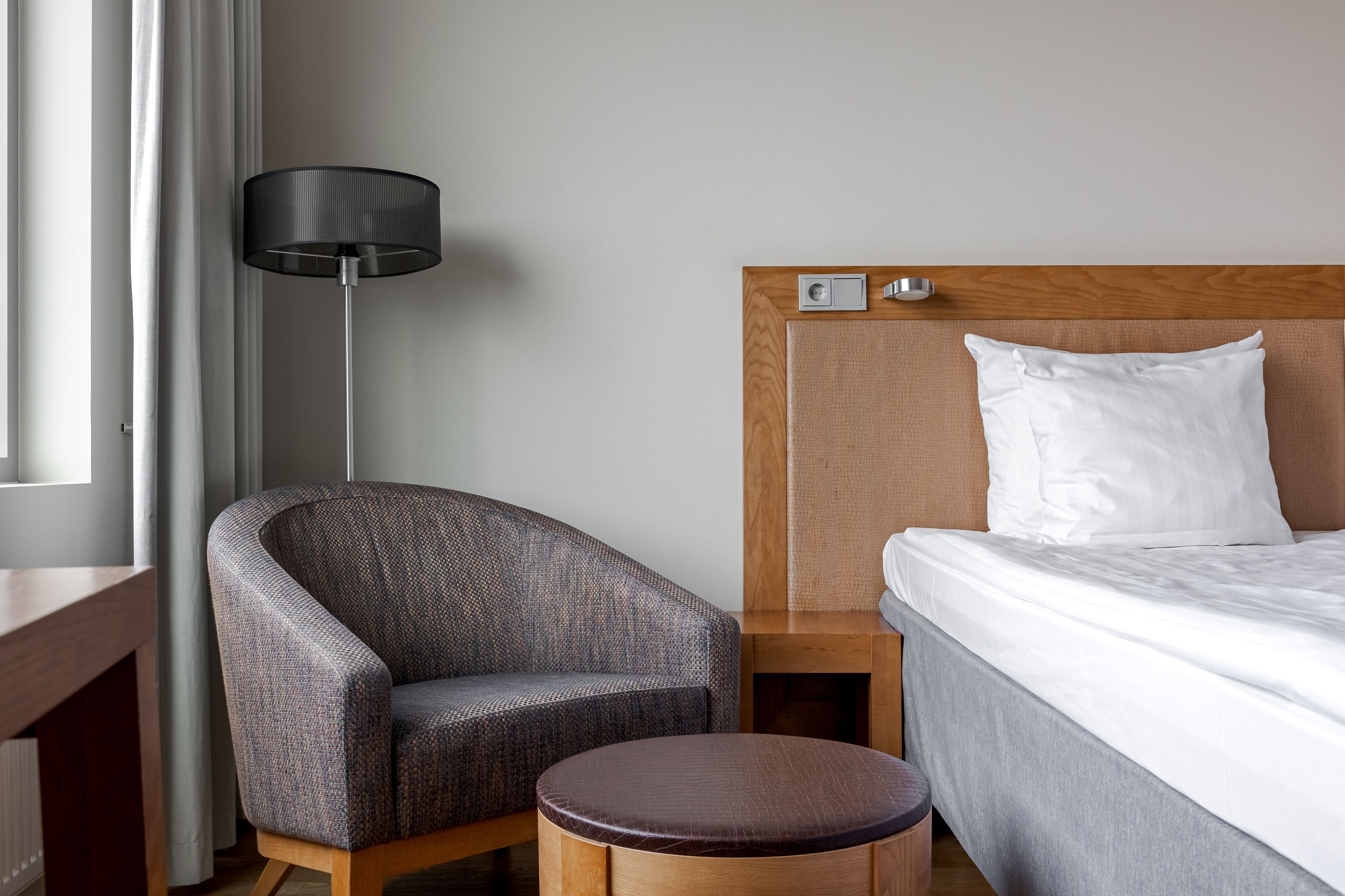 Habitación de hotel con un sillón gris curvo, un taburete de madera redondo, una cama con ropa blanca, una mesita de noche de madera, una lámpara de pie negra y una ventana con cortinas.
