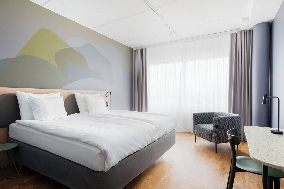 A modern hotel room with a large bed, a small table, a chair, and a view of the city through the window.