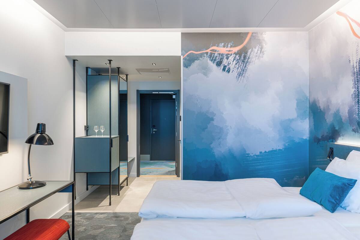 A modern hotel room with a large bed, a desk, a chair, a mirror, and a bathroom visible through an open door.