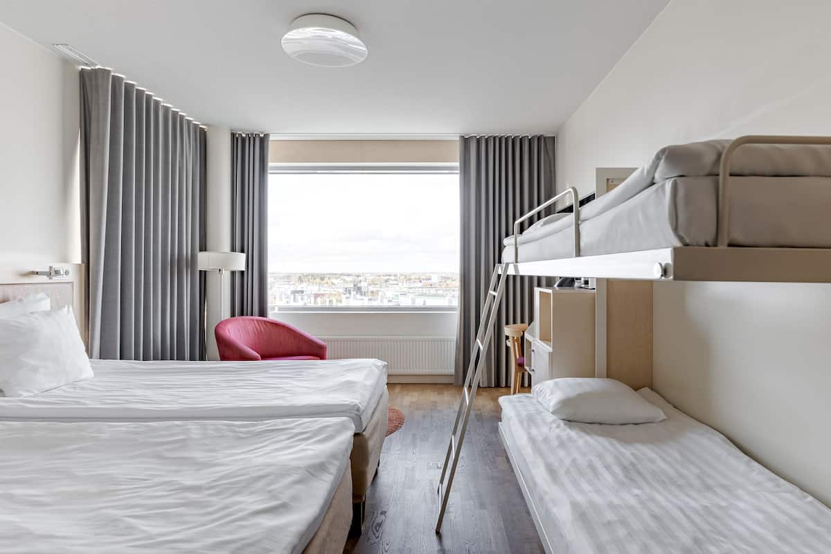 A hotel room with two beds, a red chair, a window with a city view, and a bunk bed.