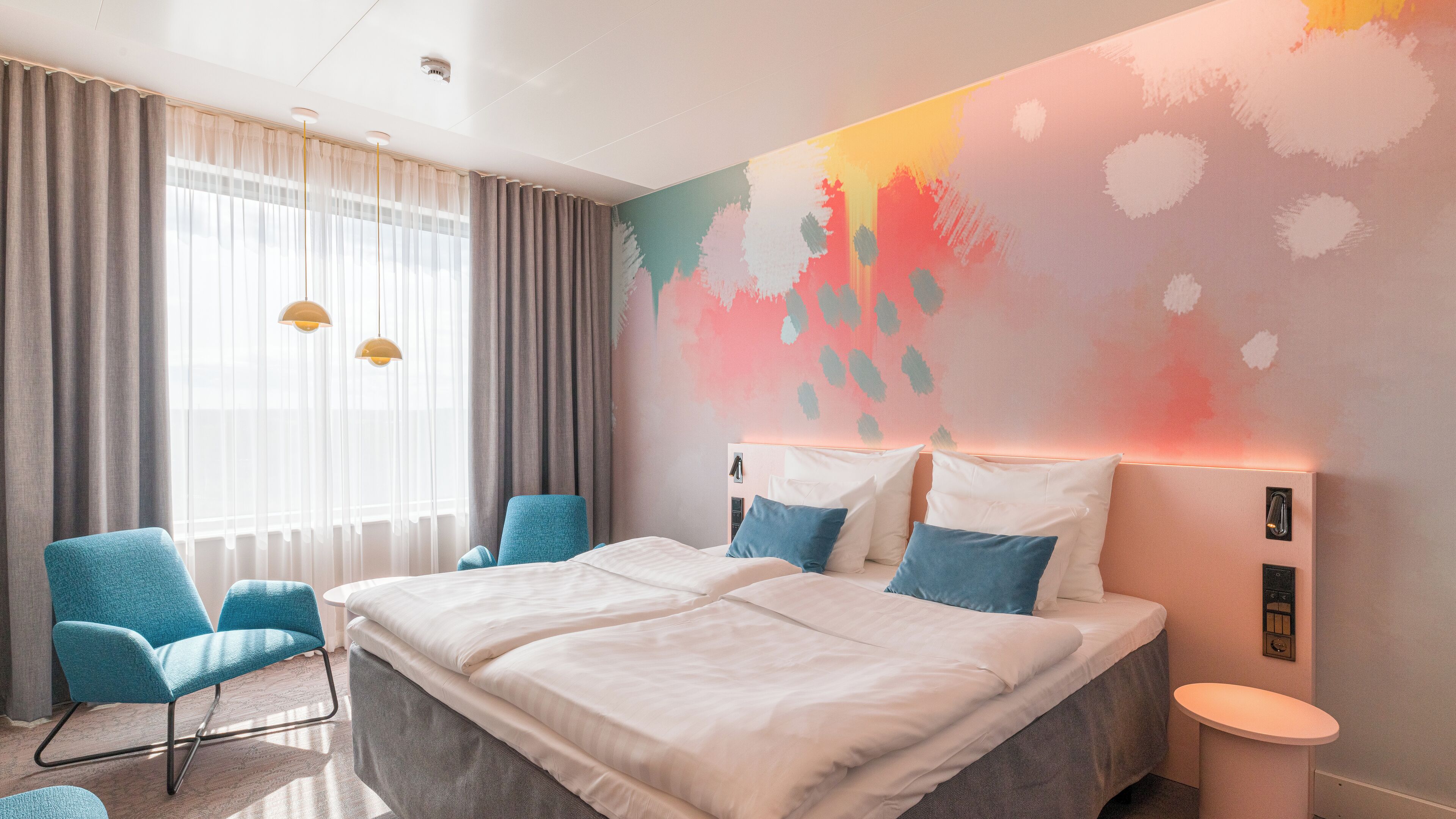 A modern hotel room with a large bed, two blue chairs, a small round table, and a colorful abstract wall mural.