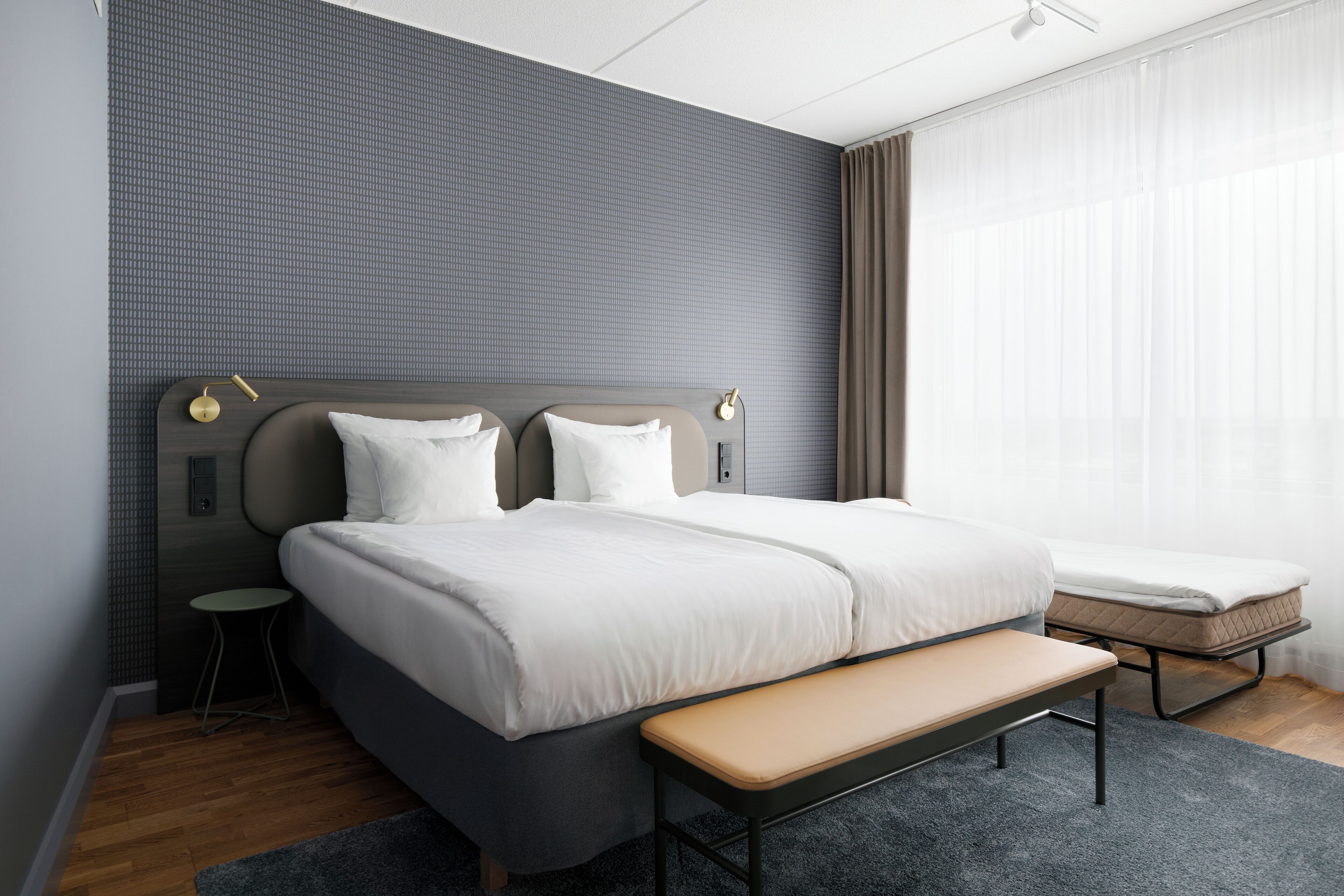 Ett modernt hotellrum med en stor säng, en bänk, ett litet bord och ett fönster med gardiner.
