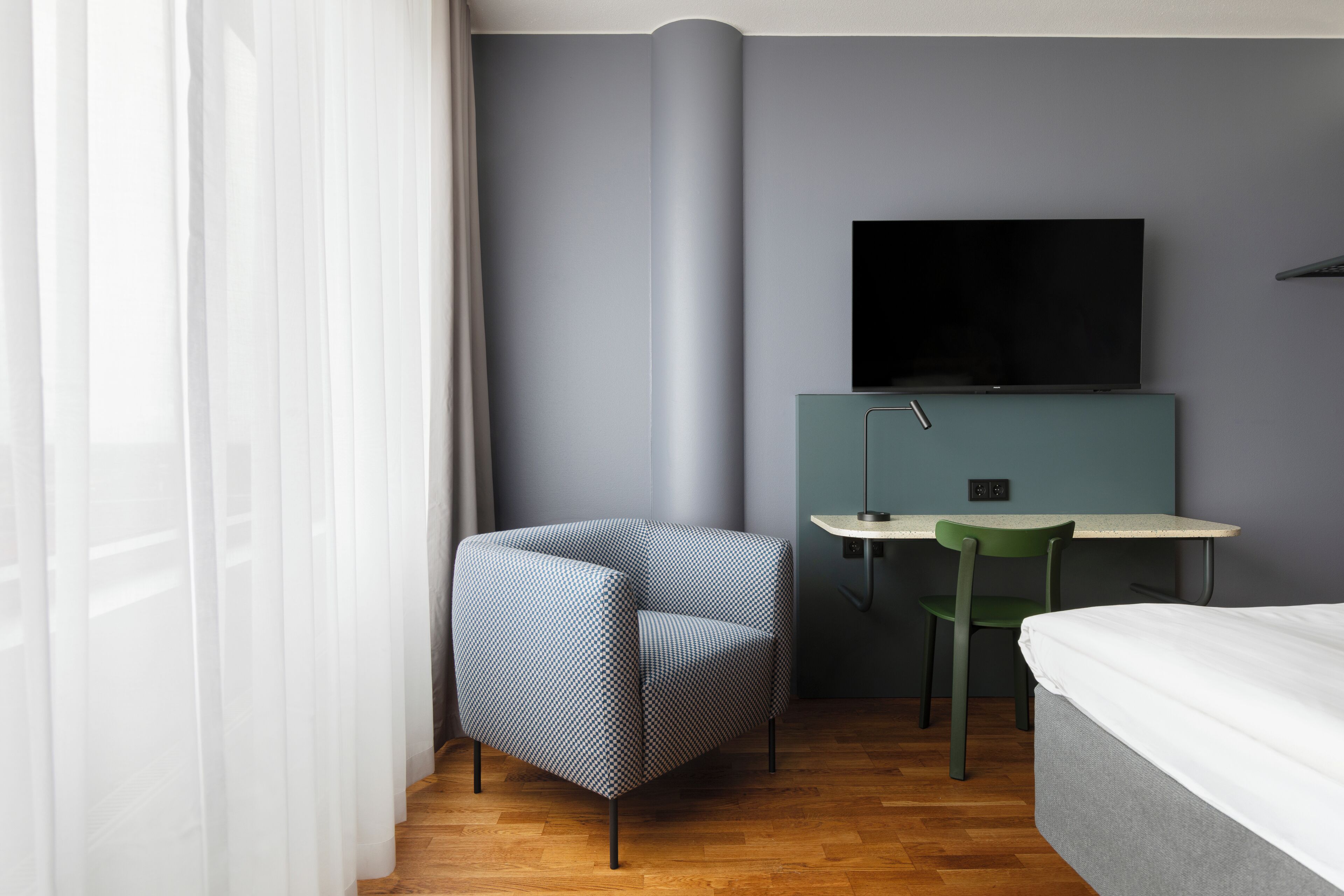 A modern hotel room with a grey wall, a flat-screen TV mounted above a teal desk, a patterned armchair, and a bed with white bedding.