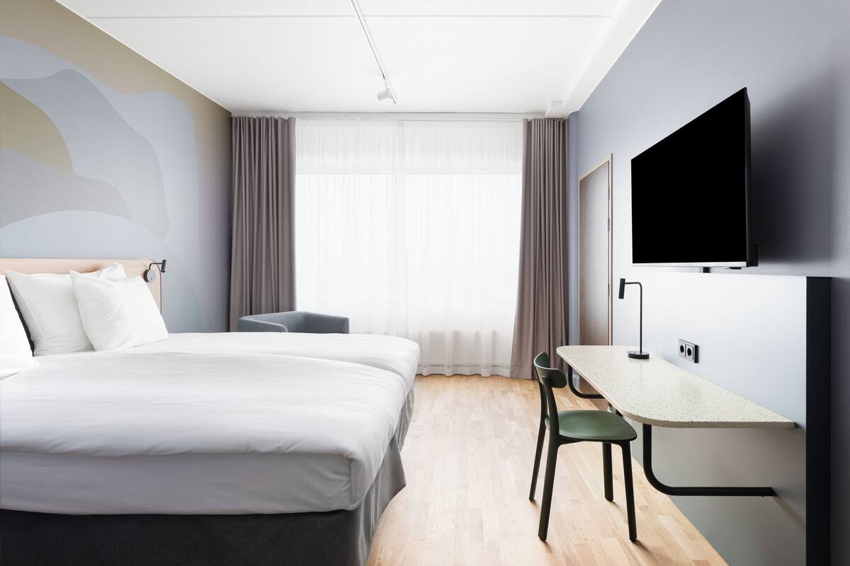 A hotel room with a bed, a desk with a chair, a TV, and a window with curtains.