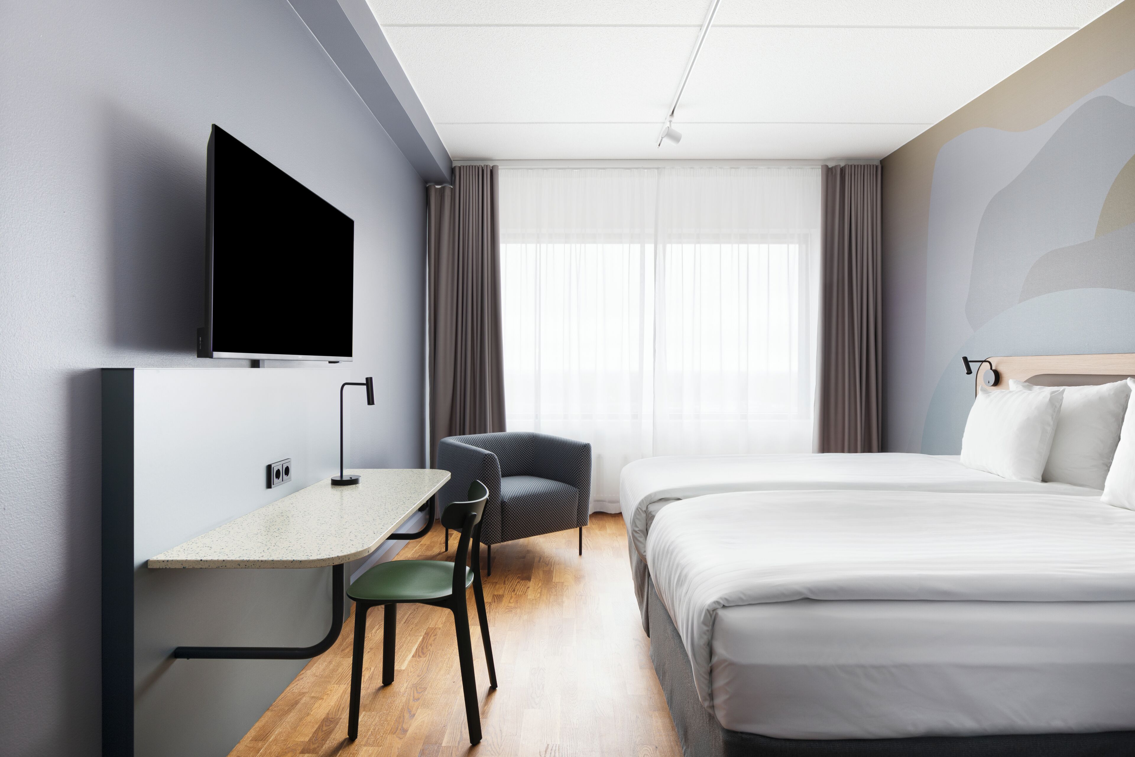 Et moderne hotelværelse med et fladskærms-tv på væggen, et skrivebord med stol, en seng med hvide sengetøj og et stort vindue med gardiner.