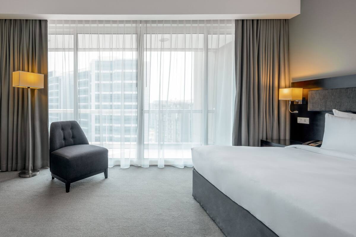 Een hotelkamer met een groot bed, een stoel en uitzicht op een stadsgezicht door dunne gordijnen.