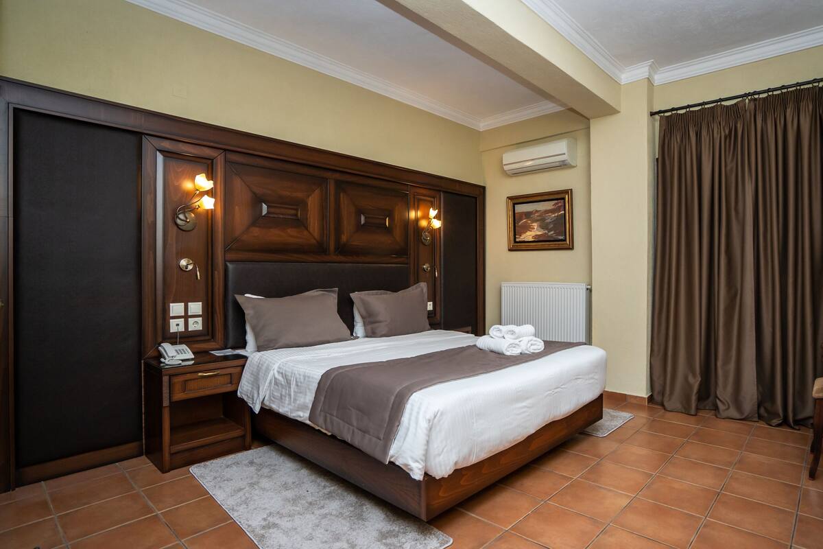 A hotel room with a large bed, wooden headboard, a bedside table with a lamp, a radiator, and a chair.