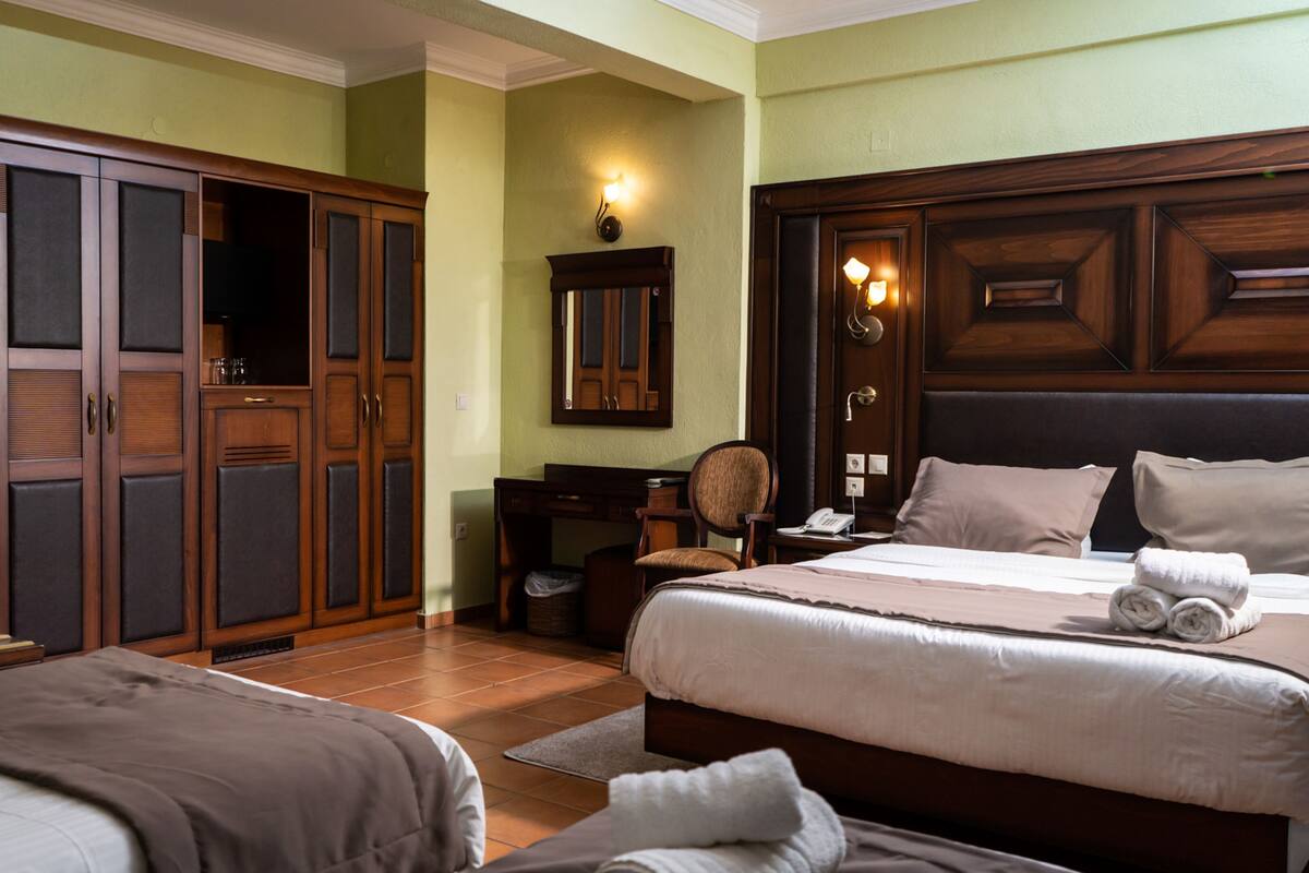 A hotel room with a large wardrobe, a desk with a chair, a bed with two pillows, and a nightstand with a lamp.