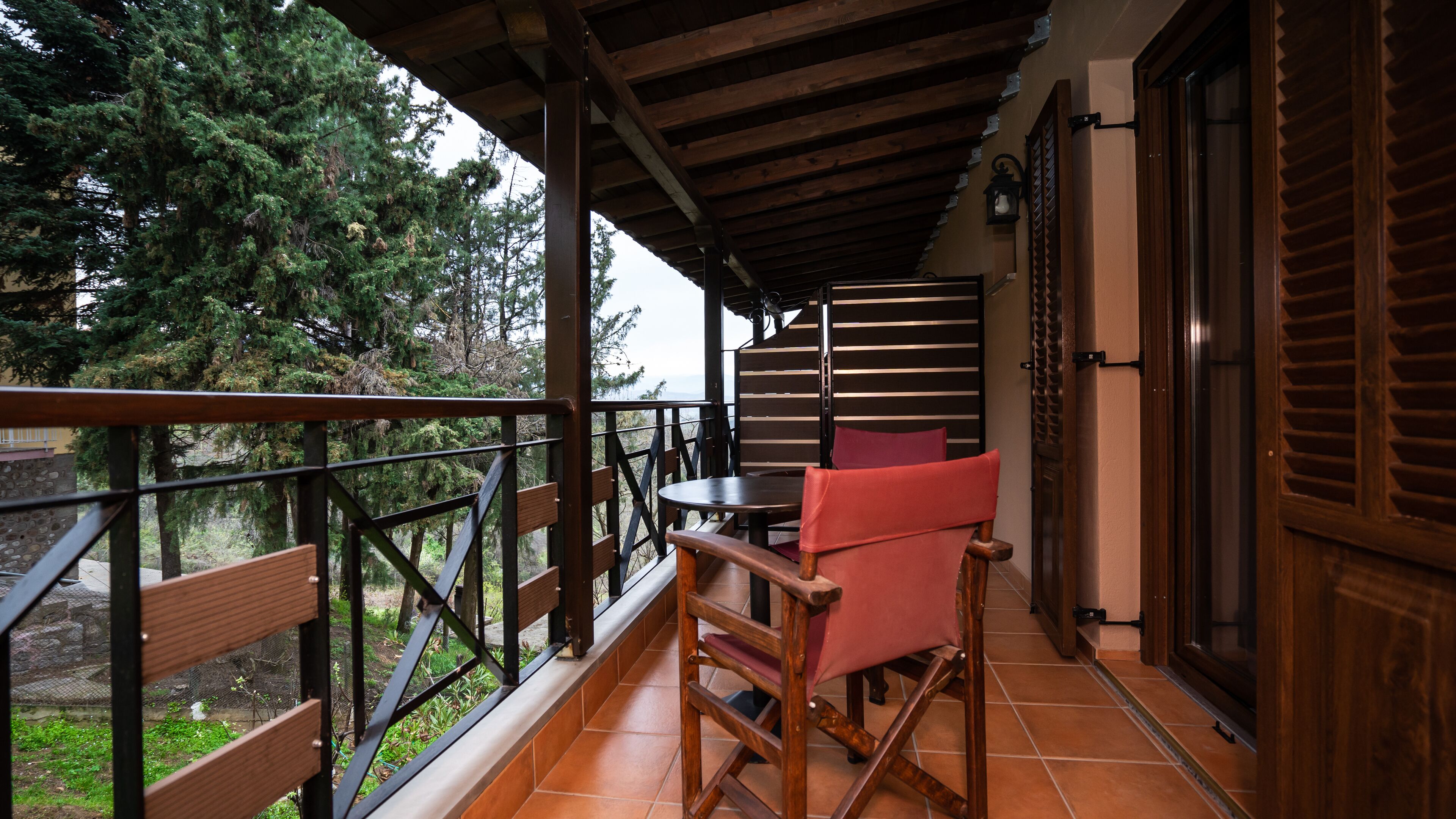 A balcony with a wooden table and red chairs, a view of trees, and a wooden door.