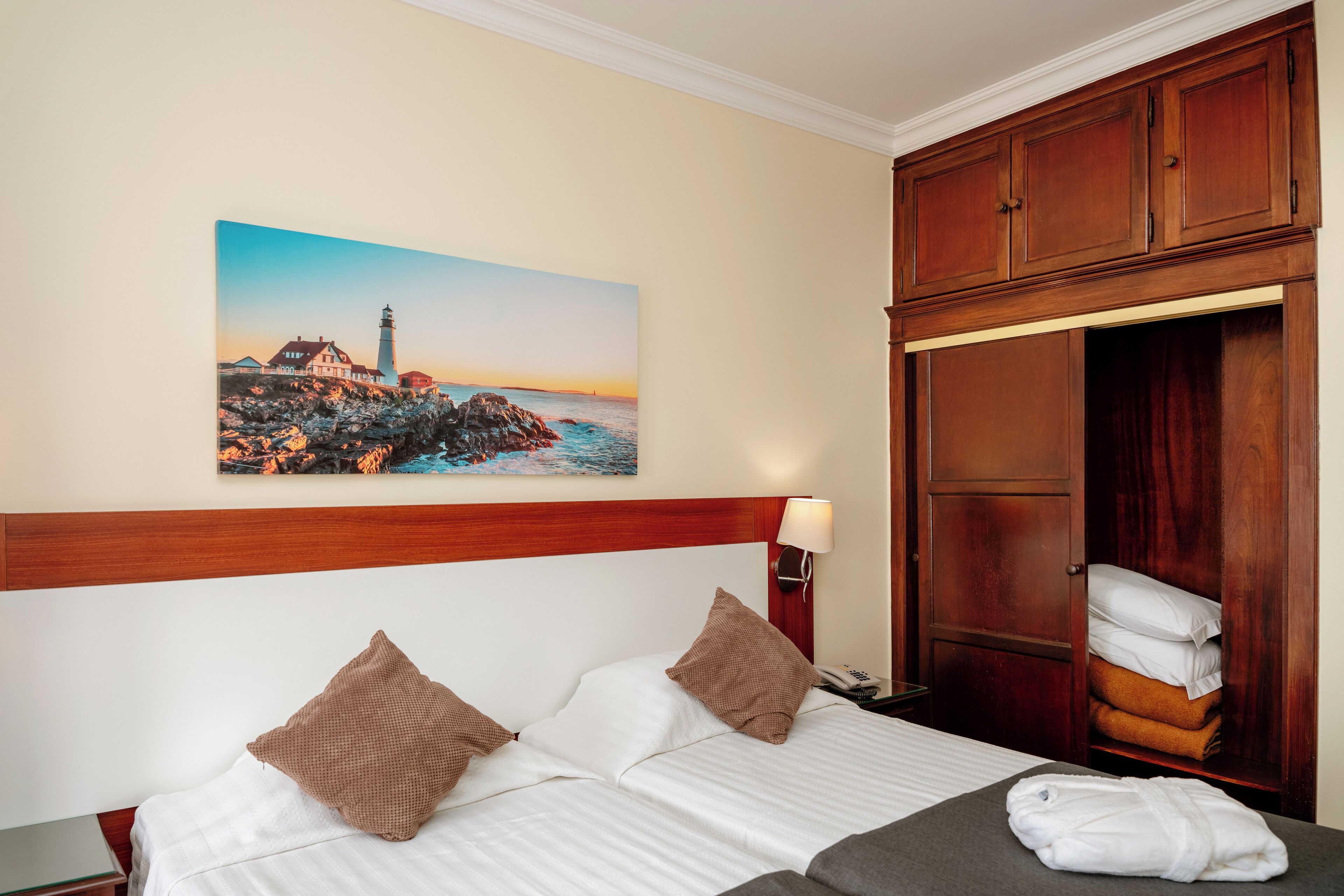 Une chambre d’hôtel avec un lit, des oreillers, un tableau représentant un phare, une armoire et une lampe de chevet.