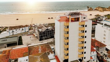 Um edifício costeiro com terraço no telhado, próximo a uma praia de areia e falésias rochosas.