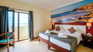 A hotel room with two beds, a chair, a painting of a beach, and a view of the sea through a sliding door. - Hotel Avenida Praia (Portimão)