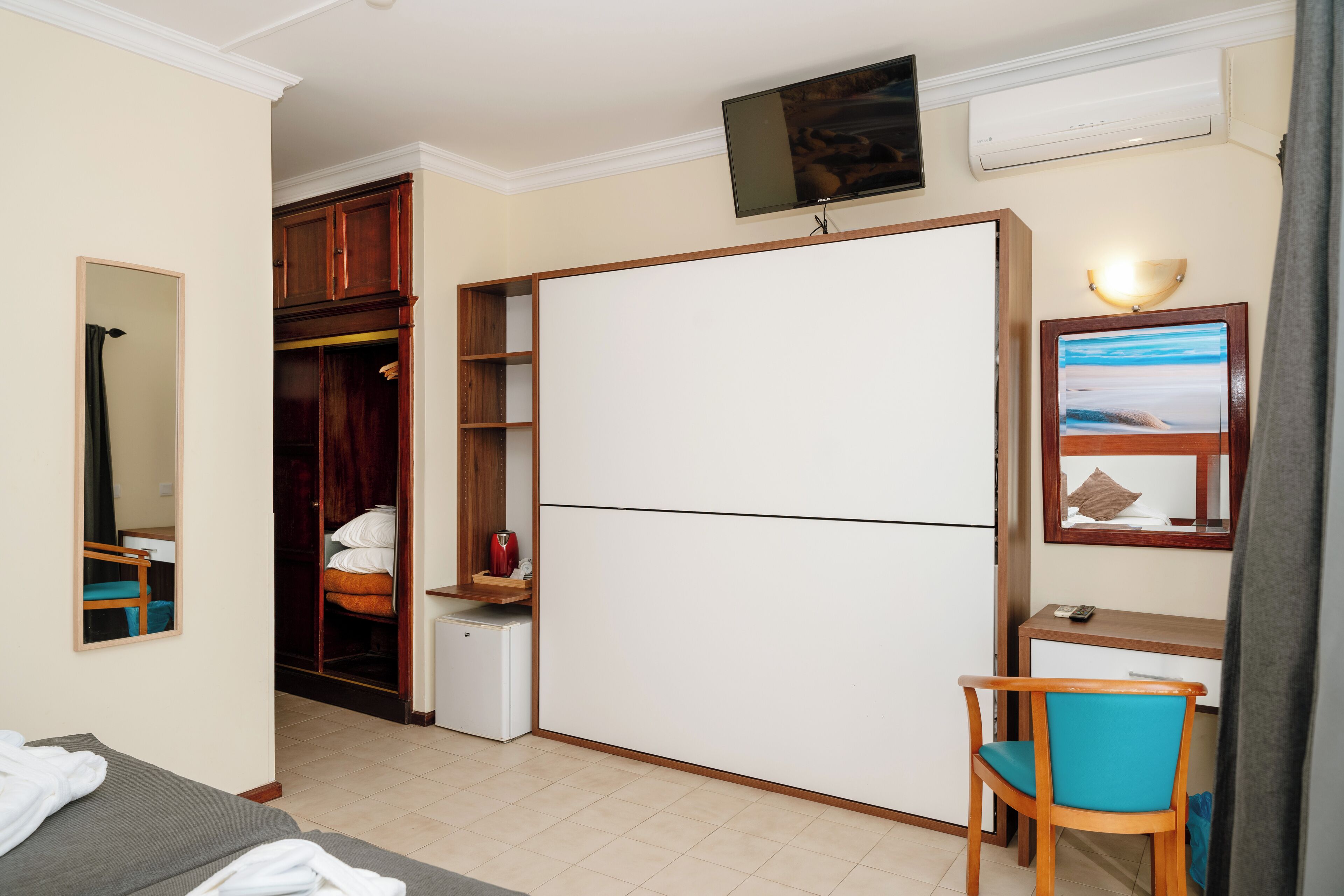 A compact hotel room with a bed, a desk, a chair, a wardrobe, a small refrigerator, and a TV mounted on the wall.