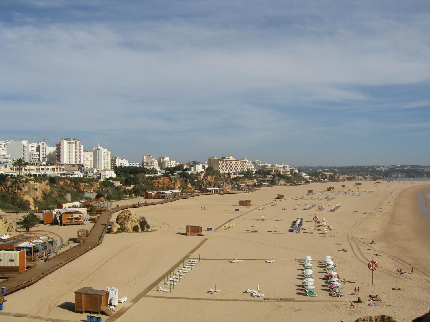 Une plage bordée d’une rangée de transats et de parasols, une étendue de sable et des bâtiments en arrière-plan.