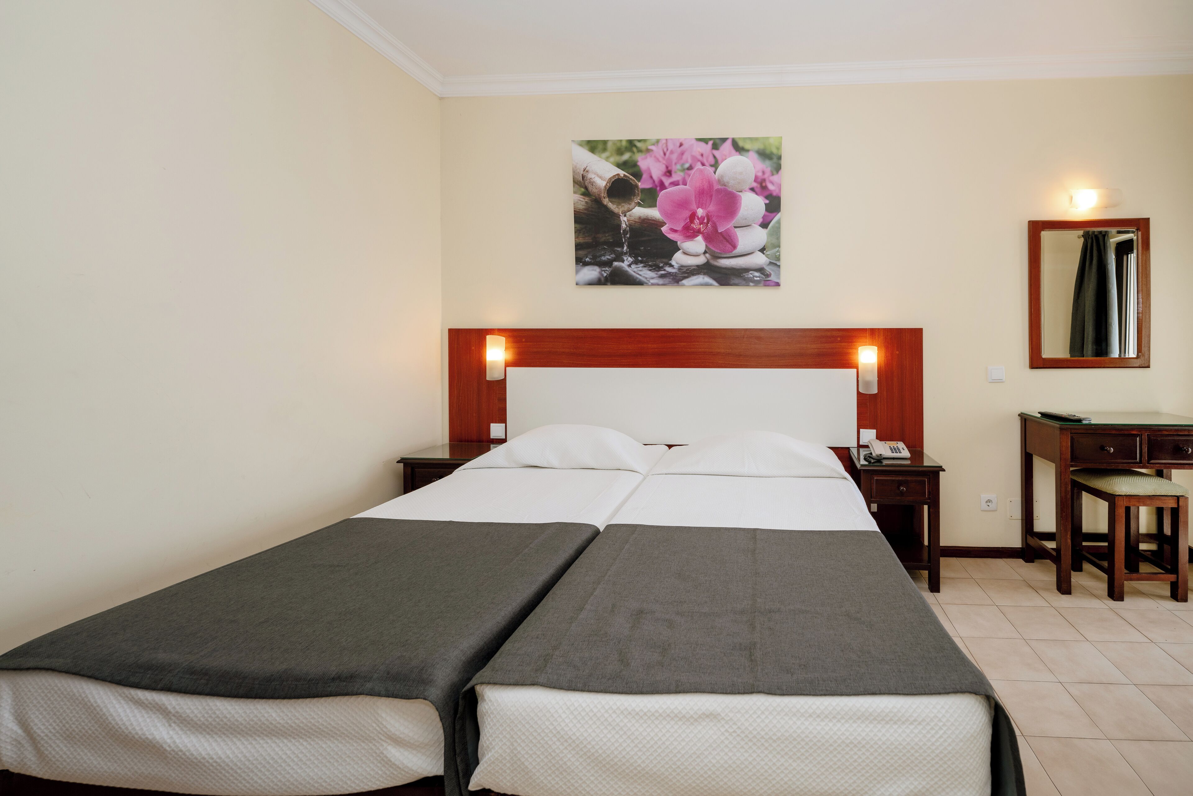 Ein Hotelzimmer mit Doppelbett, Schreibtisch, Stuhl, Nachttisch, Spiegel und einem Blumenbild an der Wand.