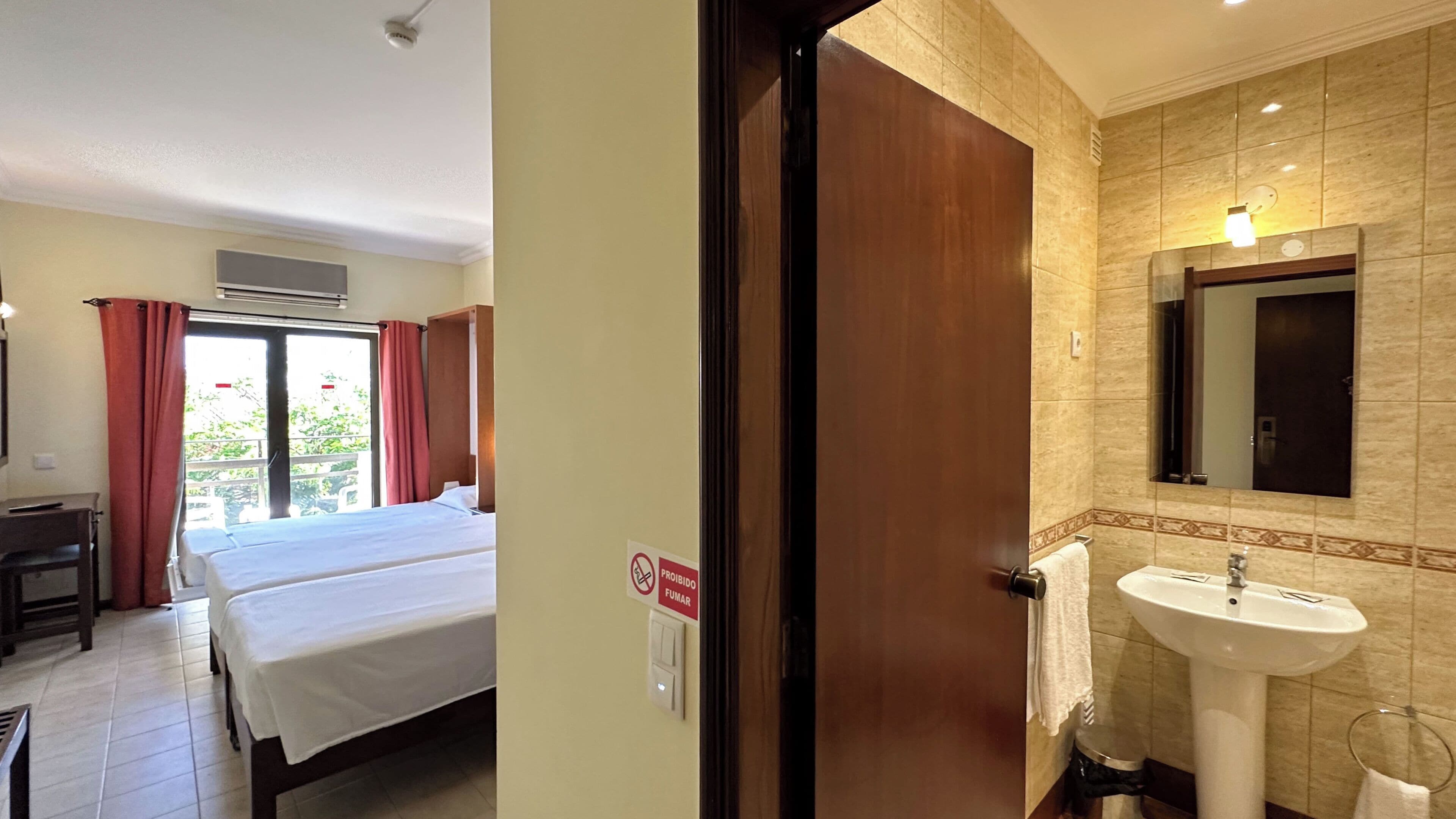 A hotel room with a bed, a desk, a bathroom with a sink and mirror, and a sign prohibiting smoking.