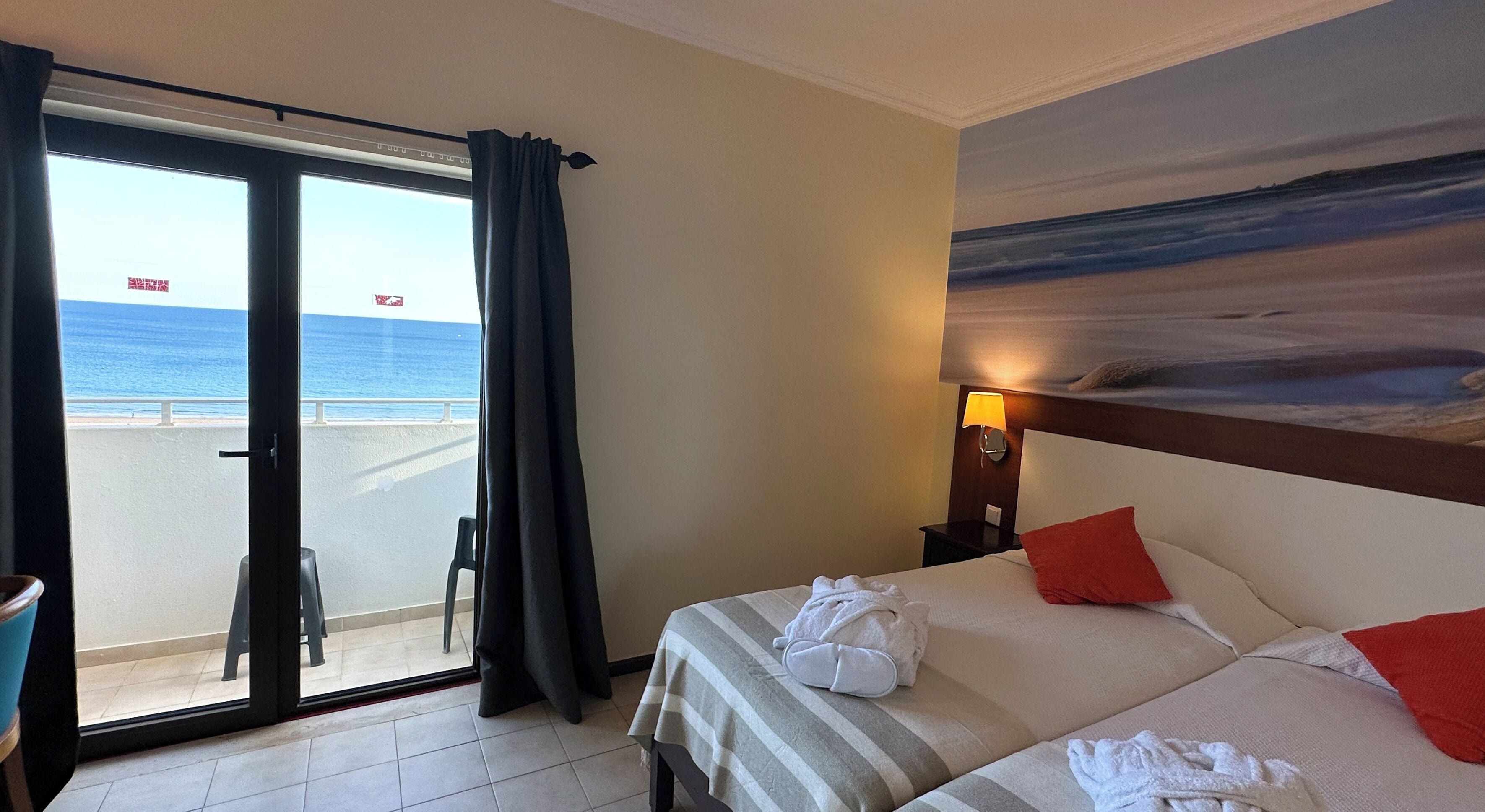 Una camera d'hotel con un letto, un comodino, un quadro raffigurante una spiaggia e vista sull'oceano attraverso una porta scorrevole.