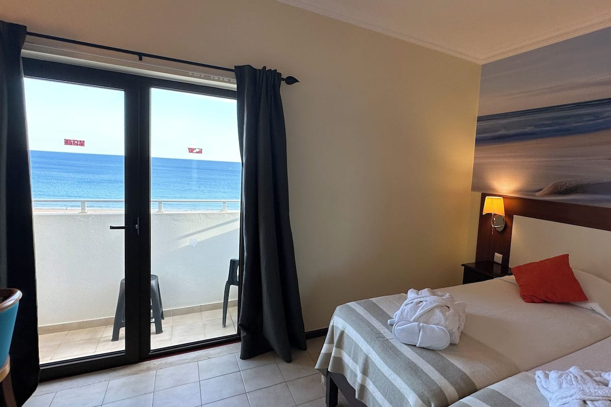 A hotel room with a bed, a nightstand, a painting of a beach, and a view of the ocean through a sliding door.